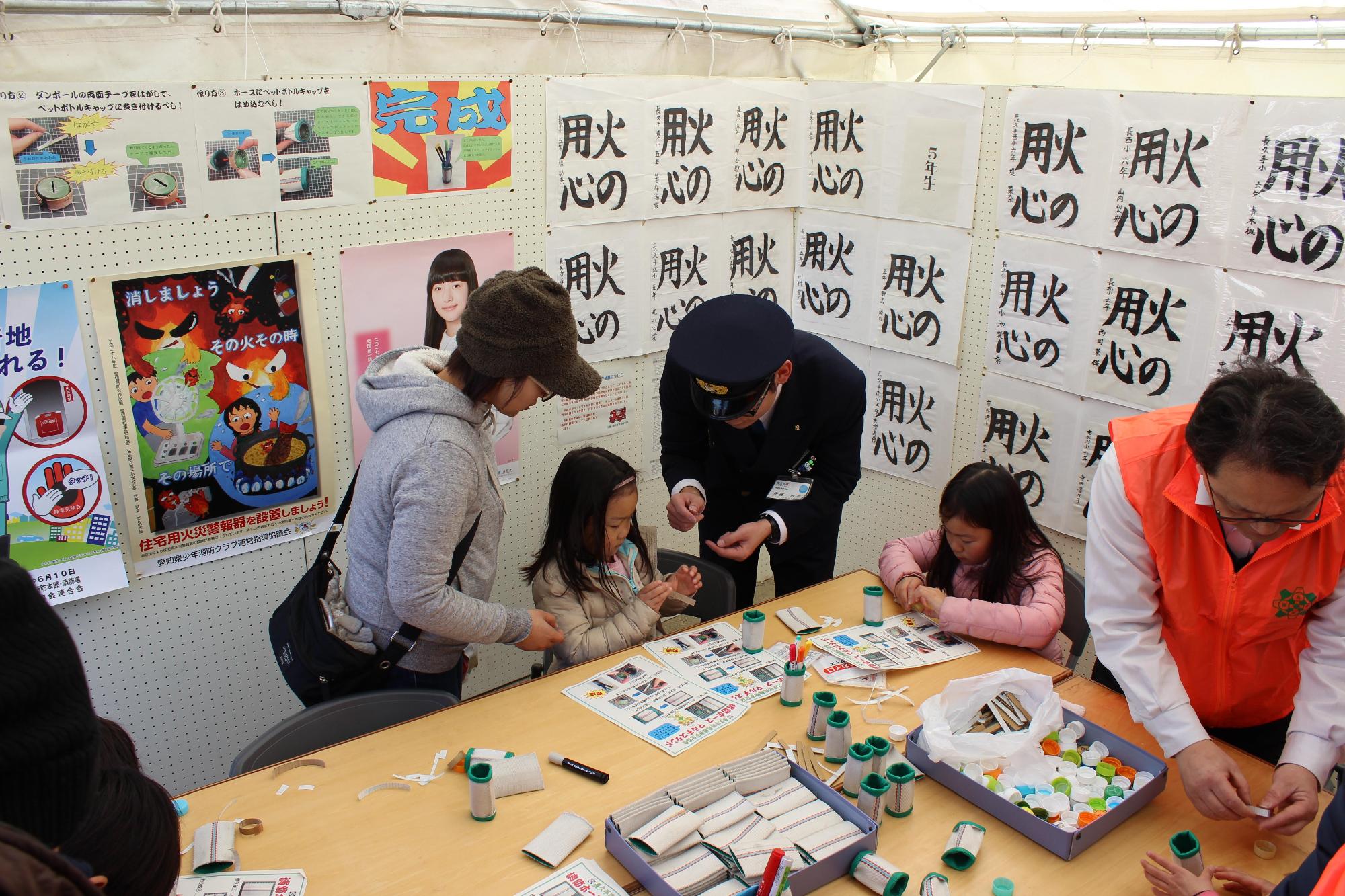 危険物安全協会ブースの壁に書やポスターが展示されており、展示コーナー前の机の上で参加者たちがマルチケースを作っている写真