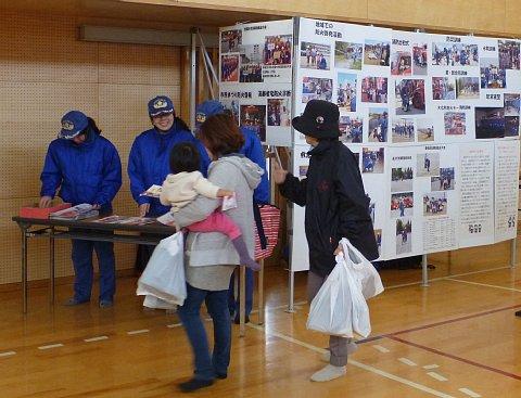 消防活動の紹介を展示しているブースの前に、青色の作業着を着た3名の女性消防団が長机の後ろに立って参加者の親子に笑顔で話している写真