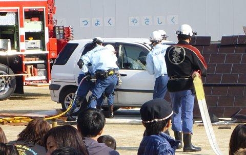 消防職員と消防団員がホースを引っ張って来て、車両からの救出訓練行っている様子を見学している参加者の写真