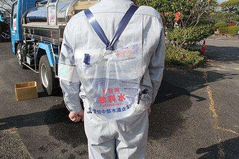 作業着を着た男性が、非常用飲料水と書かれた透明の袋を背負っている写真