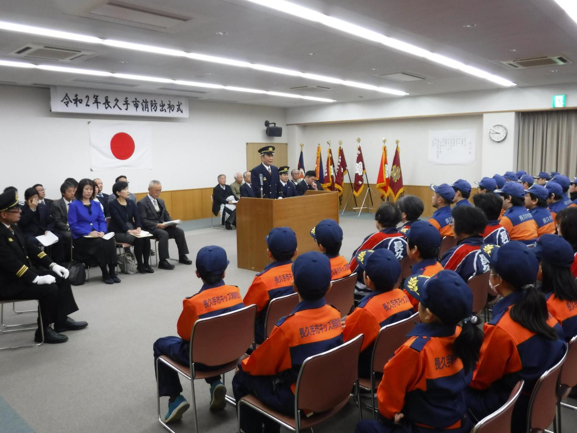 出初式にて来賓と団員が向かい合わせに座っている会場で団長が挨拶をしている写真