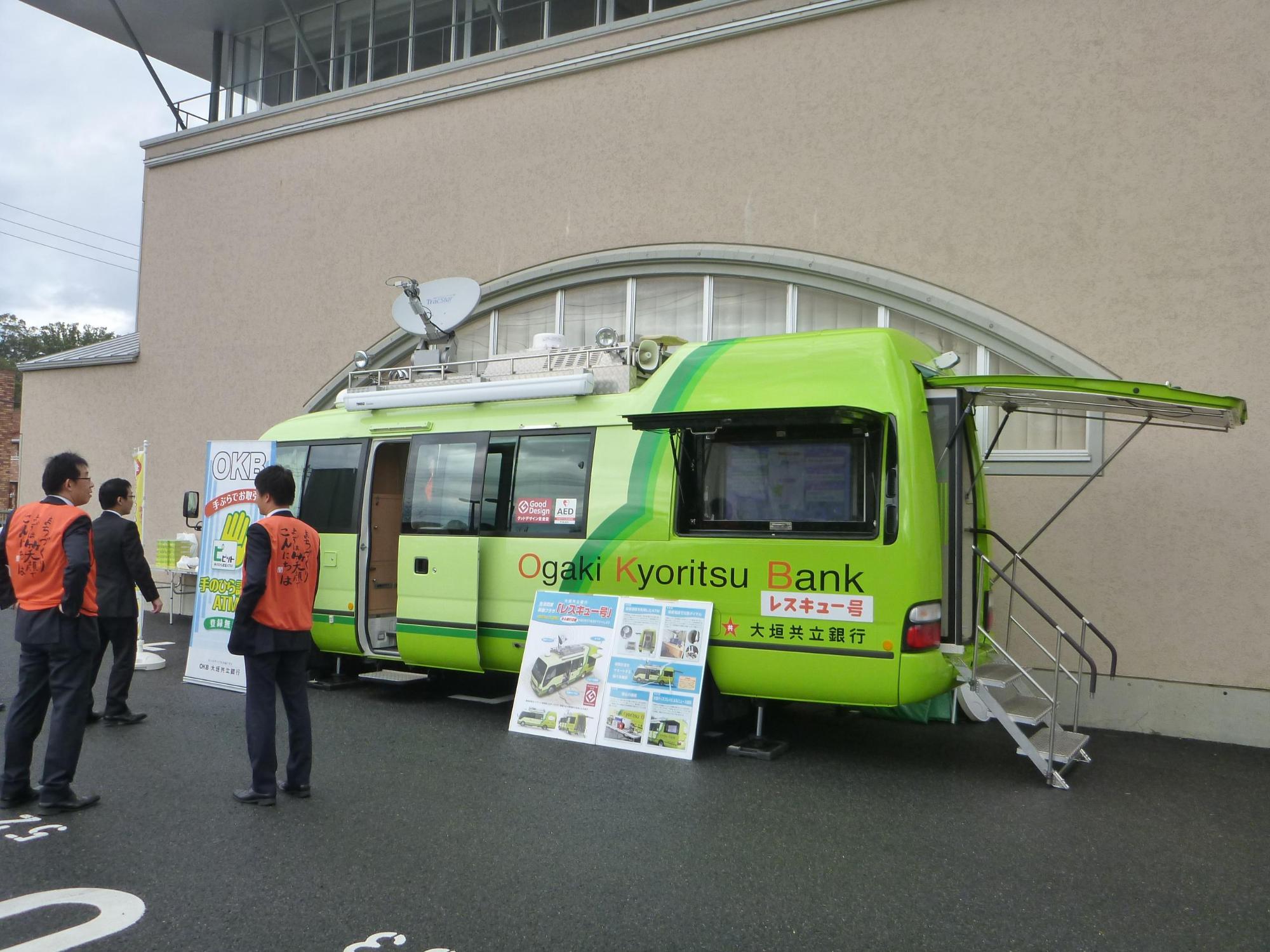 黄緑色の大垣共立銀行のレスキュー号の前にパネルが展示してあり、オレンジ色のジャンバーを着たスタッフたちが車の方を向いて話している写真