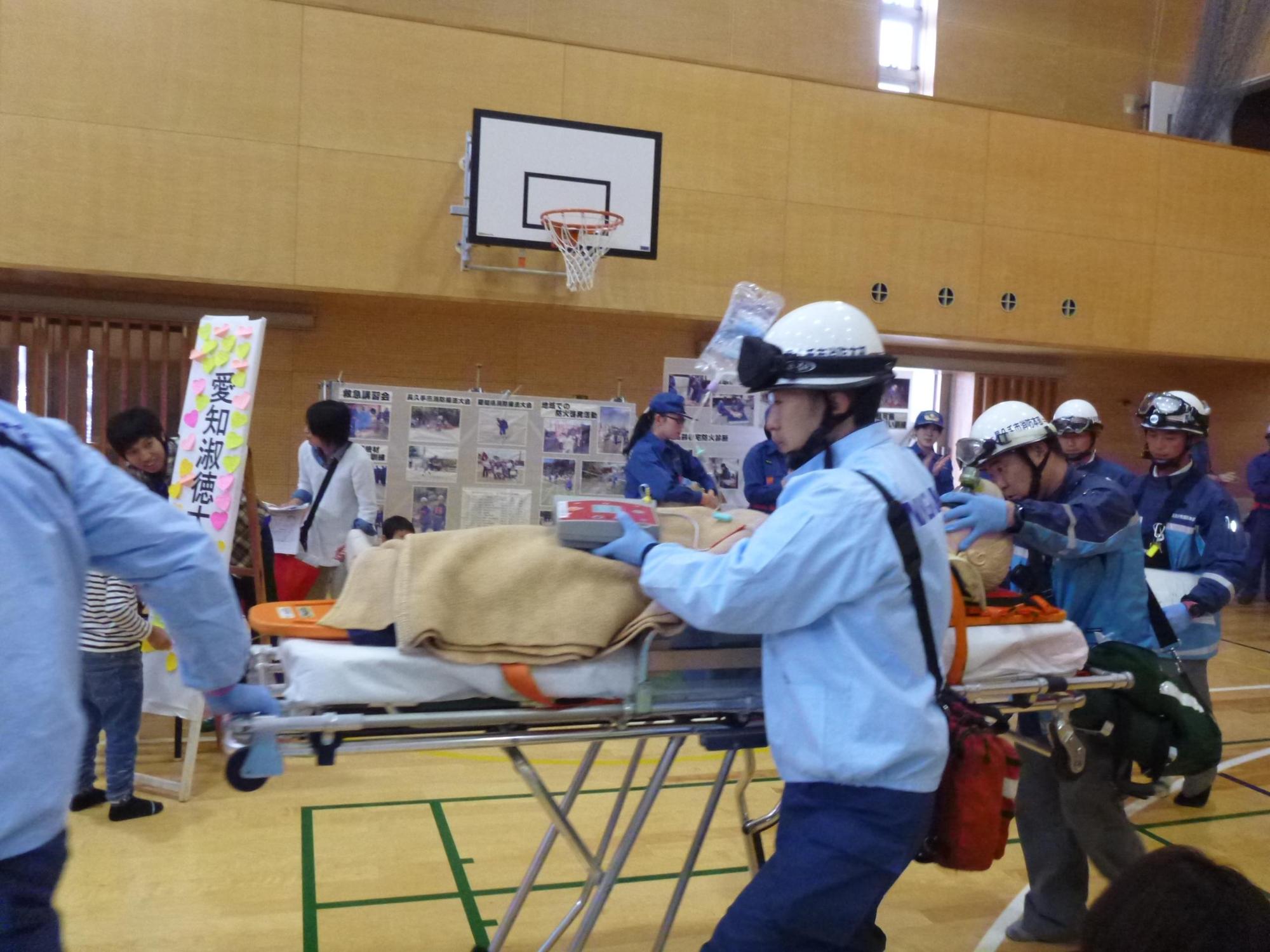 体育館で消防署員が救急活動訓練で担架に乗った人形を運んでいる写真