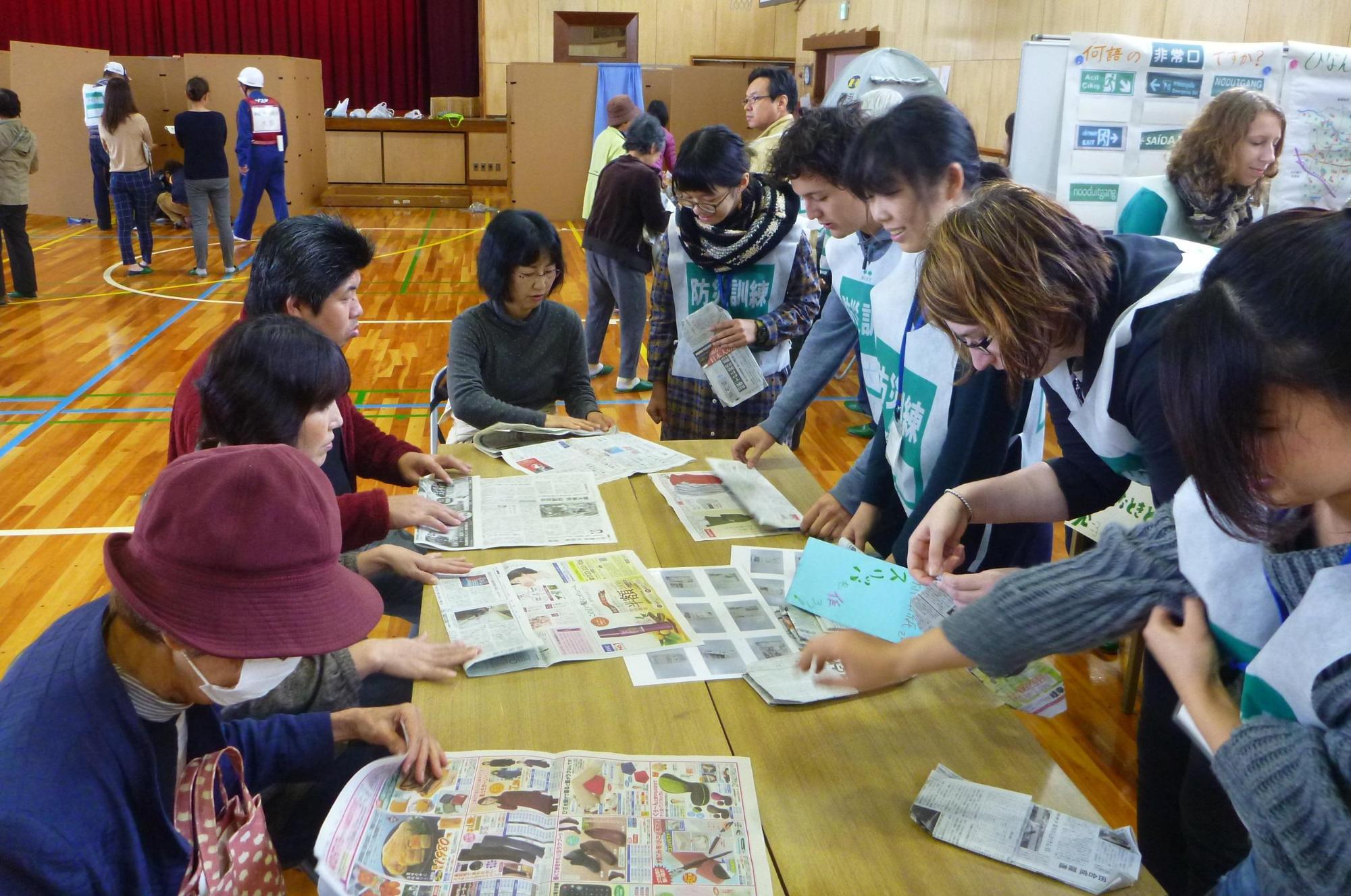 体育館に設置されたテーブルの上で、スタッフに教わりながら新聞紙を使った防災用のスリッパを作る参加者たちの写真