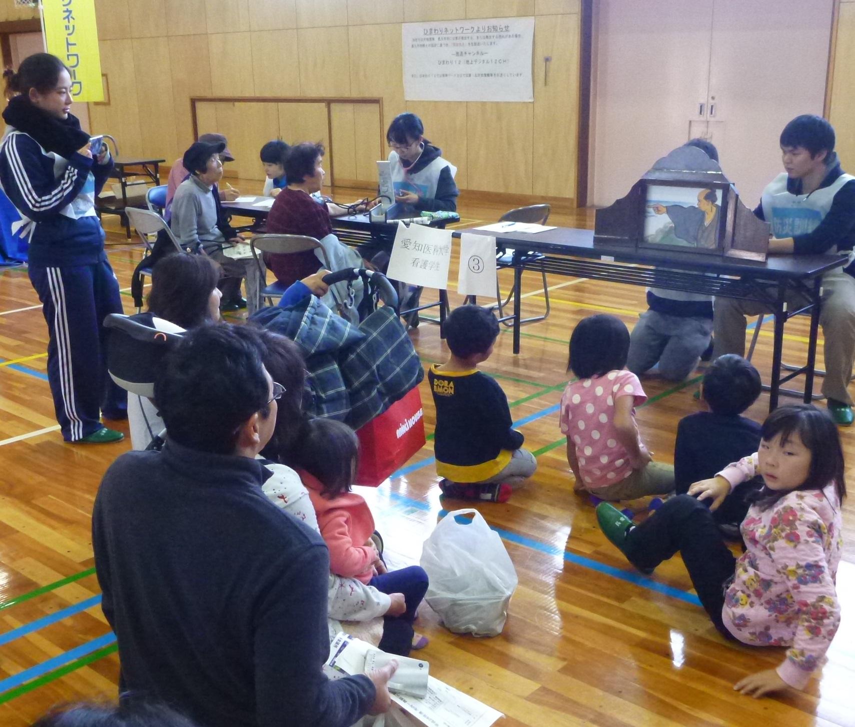 体育館に設置されたテーブルの上での紙芝居を床に座って見ている人々の写真