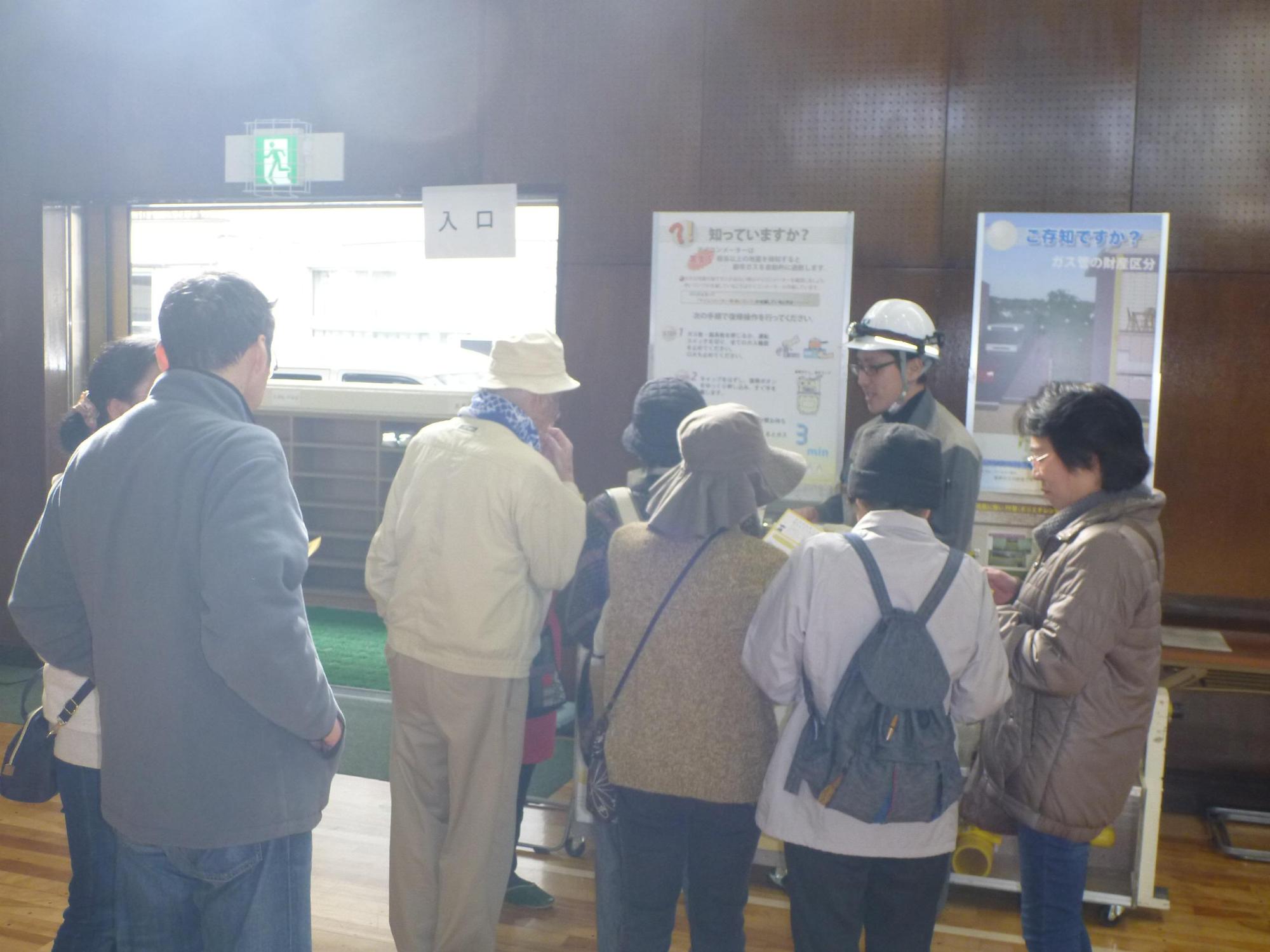 パネルが展示されている東邦ガス株式会社緊急保安センターのブースでヘルメットを被ったスタッフが参加者たちに説明をしている写真