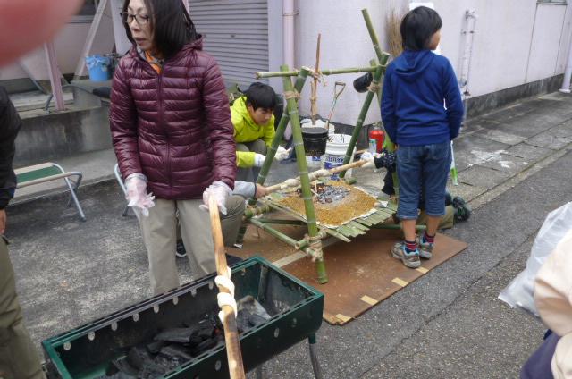ボルドー色のダウンを着た女性が棒に巻き付けたパンを炭火で焼いており、後ろの方でも黄色いジャンパーを着た男性たちが棒に巻き付けたパンを炭火で焼いているところに子供が立っている写真