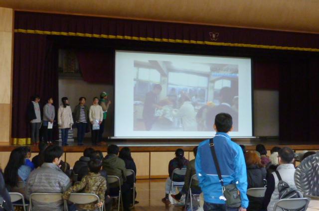 体育館の舞台のスクリーンに防災キャンプの様子が映し出され、舞台で子供たちがキャンプの成果発表している様子を椅子に座っている参加者たちが見ている写真