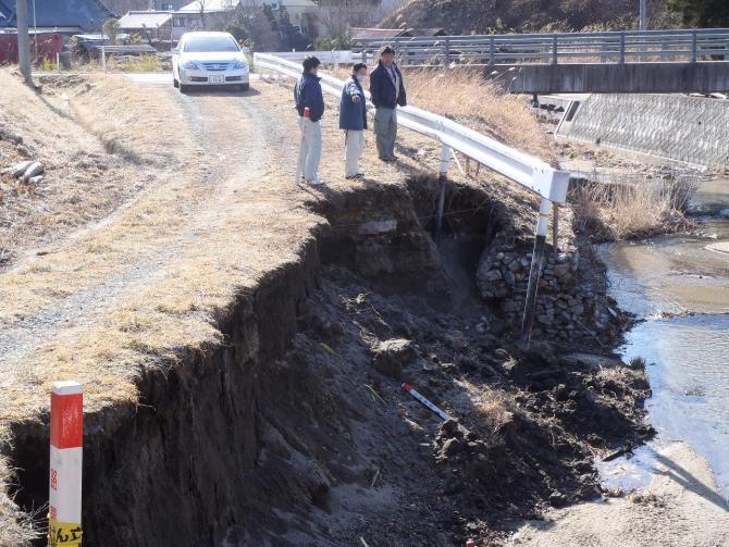 河川の左側の土手が大きく崩れており、男性3名が指差しして現地の被害の状況を確認をしている写真