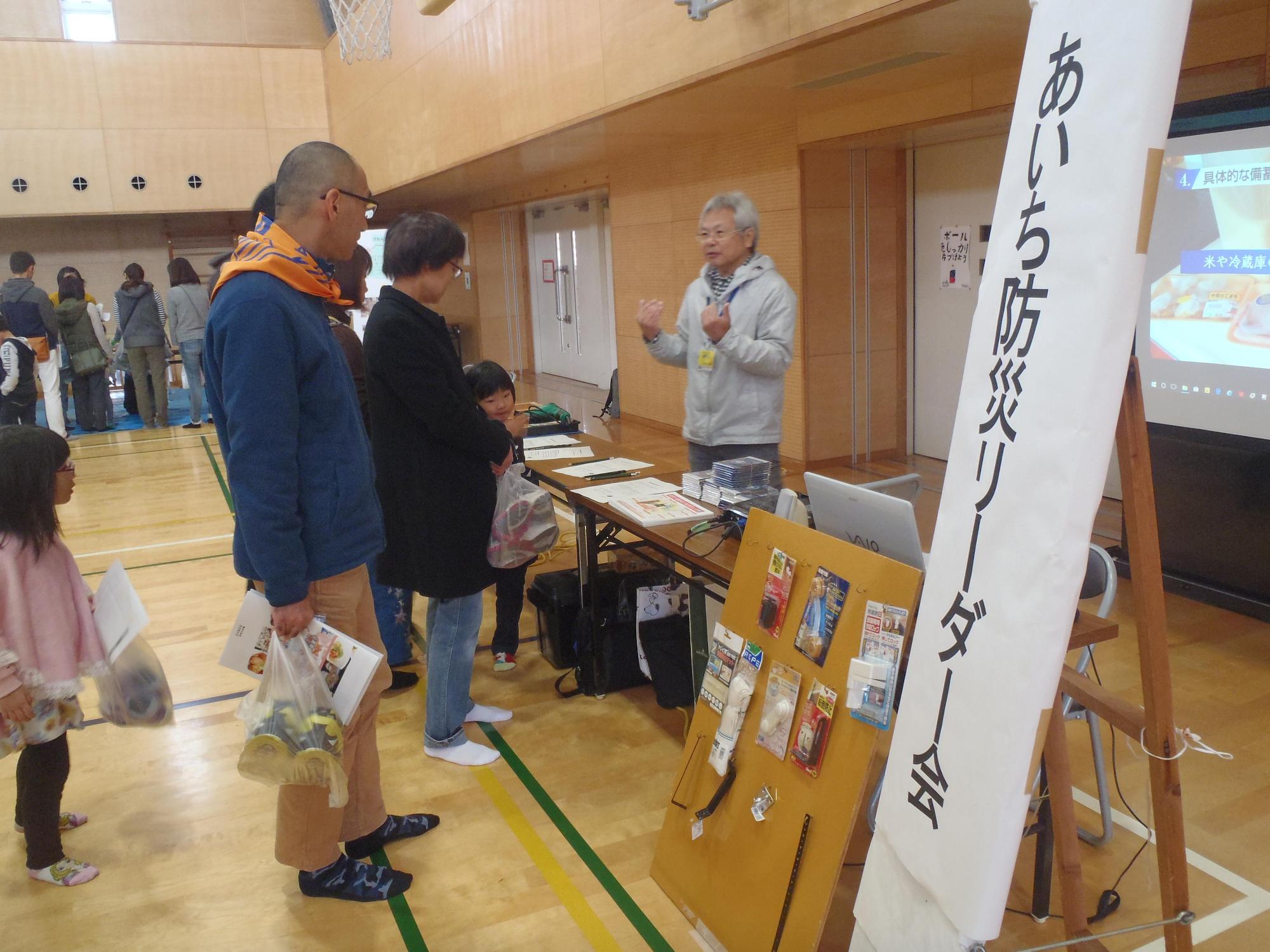 転倒防止用のグッズが展示されたあいち防災リーダー会のブースでスタッフより説明を聞く家族連れの人々の写真