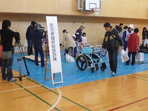 体育館の中にビニールシートを引いている上に「防災資機材取扱訓練」と書かれた看板や車いすなどが設置されており、参加者たちがそれぞれ訓練している写真