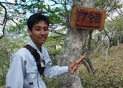 森の中で7合目と書かれた札が木にかかっており、その札を男性職員が、枝で指し示している写真(商業観光課で勤務する下谷主事)
