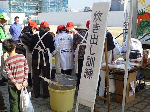 「炊き出し訓練」と書かれた看板の後ろで、オレンジ色のキャップを被った女性たちが炊き出しを行っている様子を参加者の子どもが見ている写真