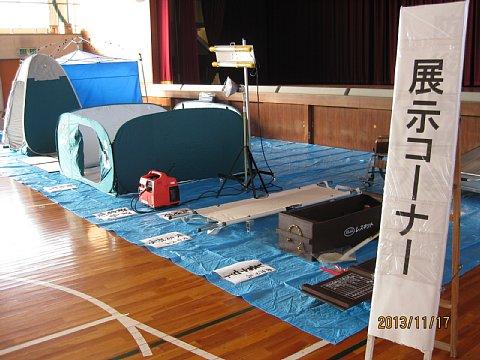 体育館のステージの下に、簡易トイレなどの防災資機材が展示されれいるコーナーの写真