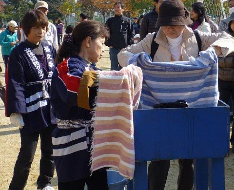 法被を着た女性消防クラブのスタッフと女性の参加者が布を持ち、天ぷら鍋の火災消火訓練している写真