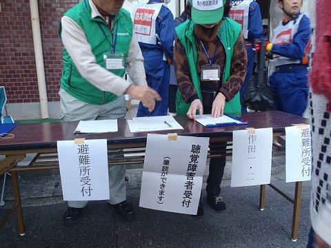 「避難所受付」と書かれた貼り紙が長机の前に貼られ、緑色のベストにキャップを被った男性と女性が受付の訓練を行っている写真