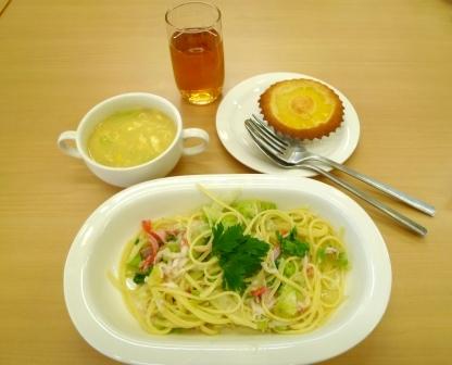 大きな白い器に盛られたスパゲッティの右上にマドレーヌとフォークとスプーン、左上にスープ、その間に飲み物が置かれた写真