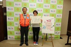 あすりーのイラストの横で感謝状をもち笑顔で記念写真をしている男女の写真
