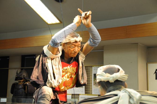 棒を手に持ち両手を頭の上に振り上げている人形の展示物の写真