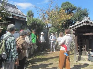 子供を抱っこした母親やリュックを背負った参加者たちが史跡(首塚)の前で話を聞いている写真