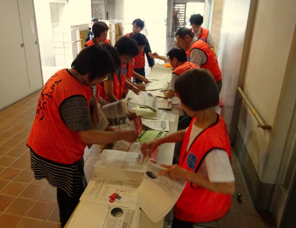 オレンジ色のベストを着たたのしみ隊の皆さんが、机の上に並べられた資料を一枚ずつ取って参加者の皆さんの為に料を準備している写真