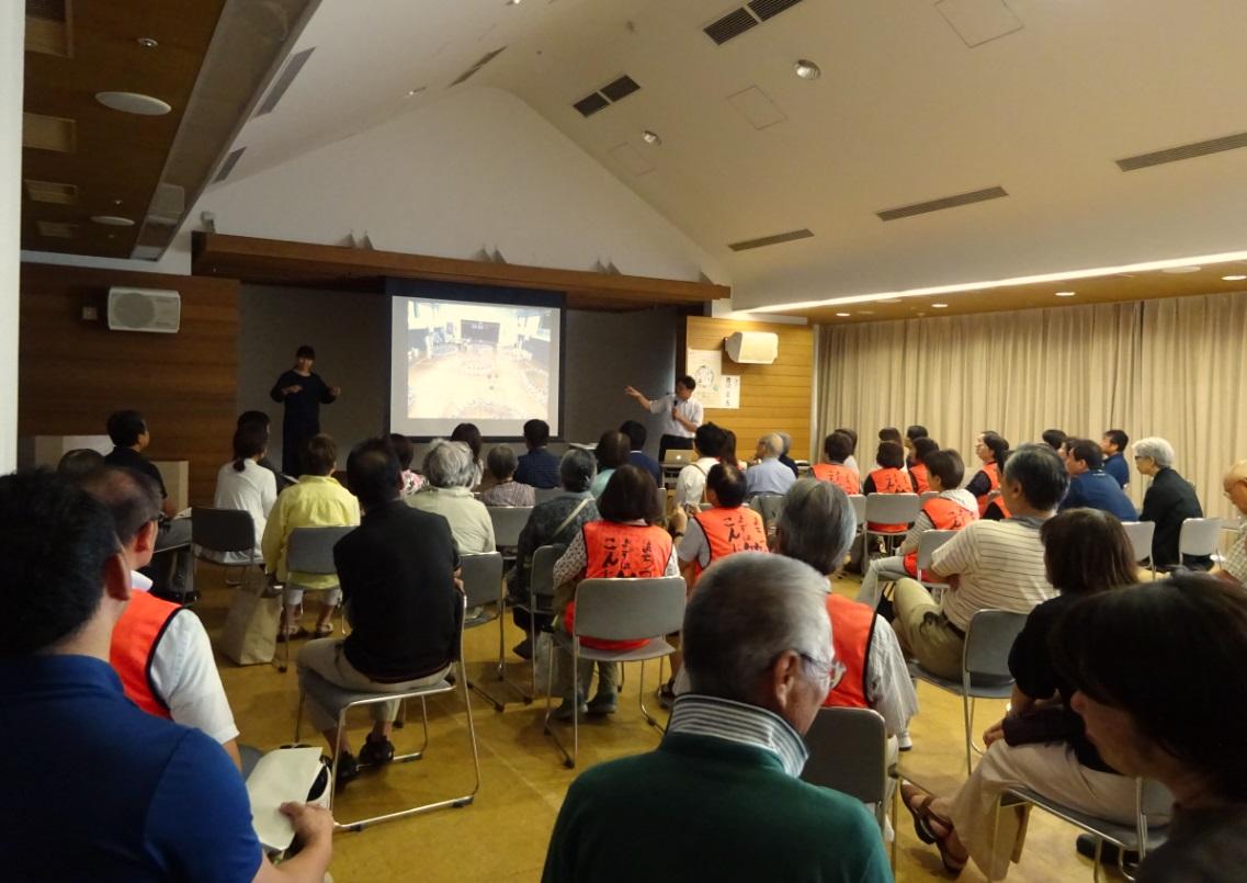 舞台上にはスクリーンに映像が映し出され、講師の方が映像を見ながらマイクを持って話をしており、その横で手話通訳者が講師の方の話を通訳しており、たくさんの参加者が椅子に座って講演を聞いている様子の写真
