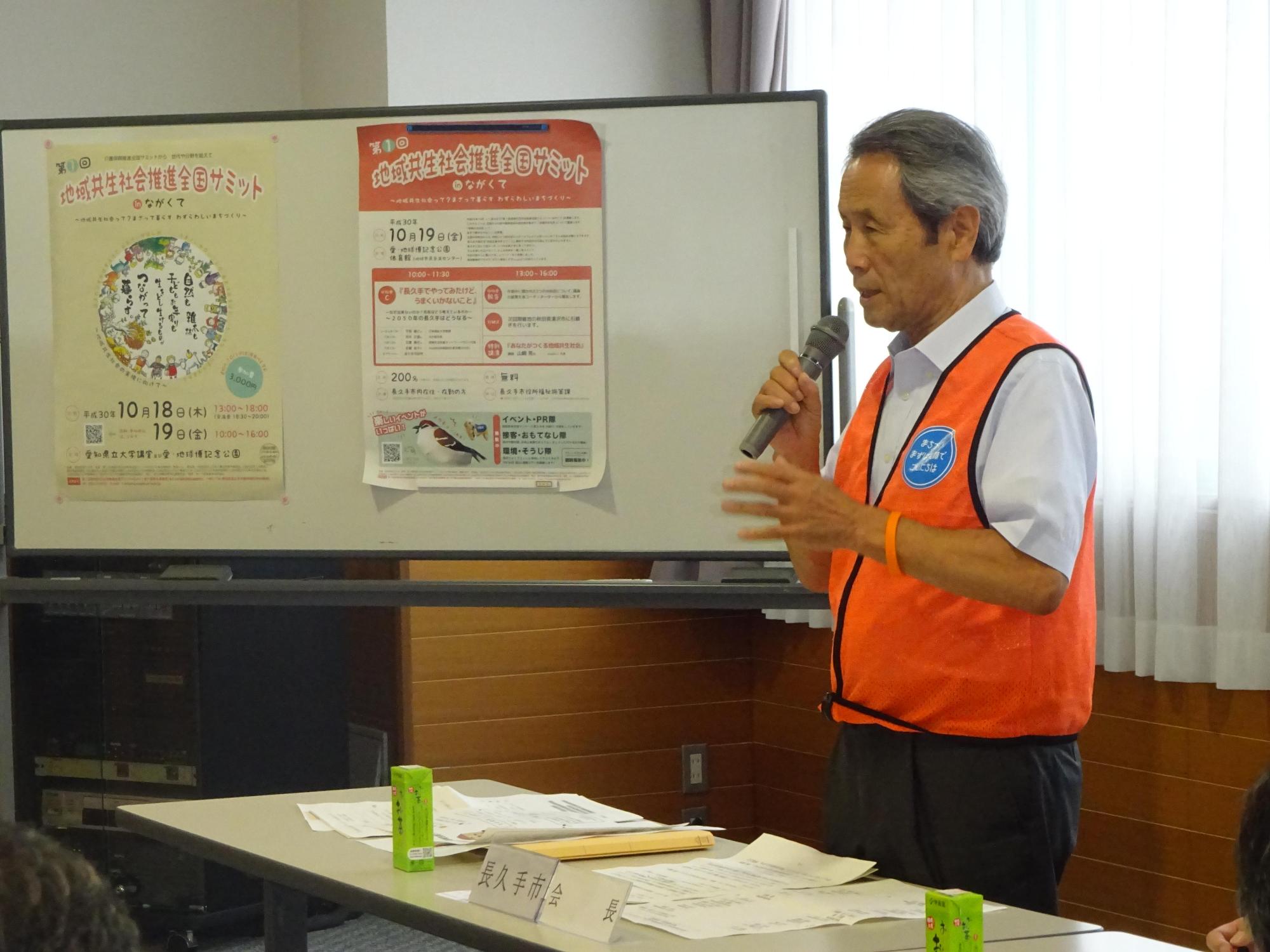 オレンジ色のベストを着た市長がマイクをもって話をしている写真