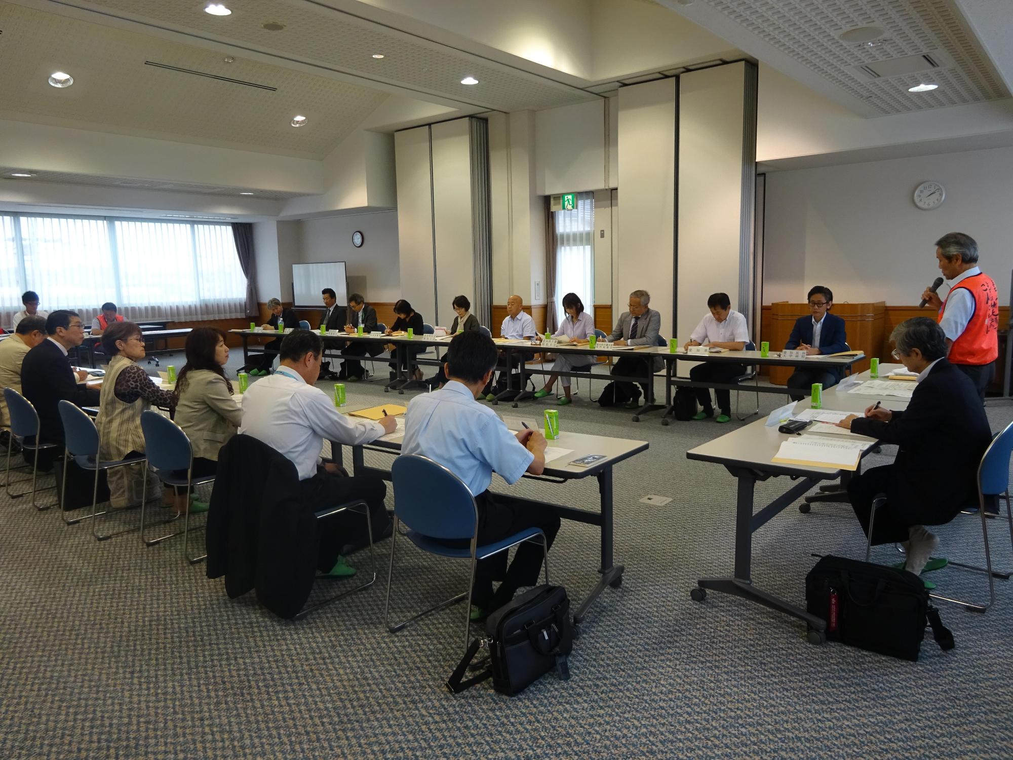コの字型に机が並んでおり、実行委員会の参加者が机の上の資料に目を通しながら、市長の話を聞いている写真