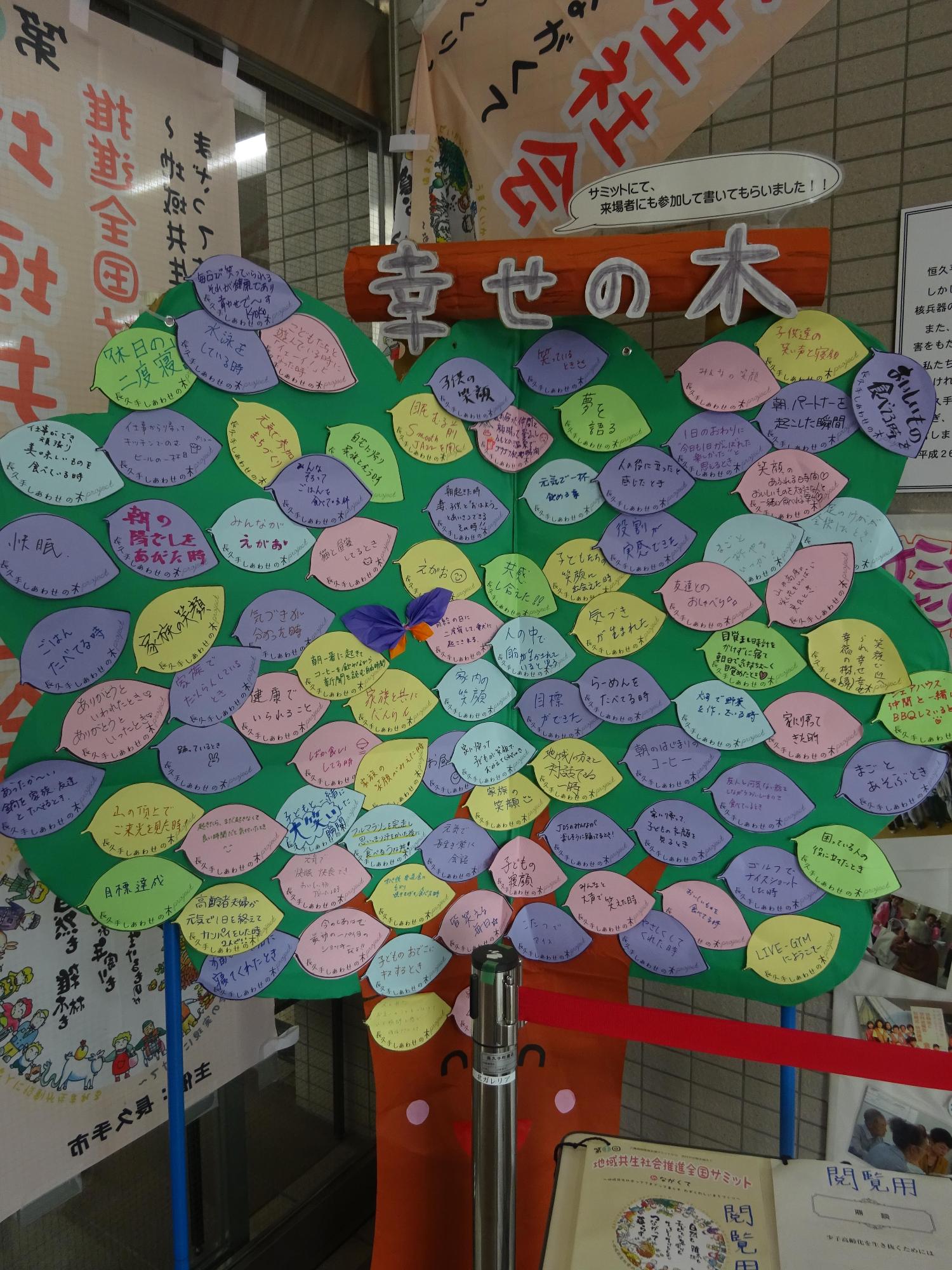 手作りの木のの幹の部分に笑顔の顔が書かれており、上の緑の部分に葉っぱをモチーフにした紫・黄緑・ピンク・黄色の葉っぱに、色々な言葉が書かれてたカードが沢山貼ってある幸せの木の写真
