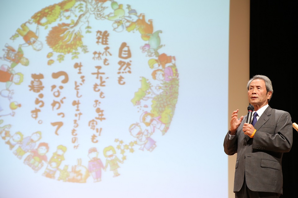 「自然も雑木林も子どももお年寄りも生きとし生けるものが繋がって暮らす」と書かれたシンボルマークが映し出されてスクリーンの前で市長がマイクを持って話をしている写真