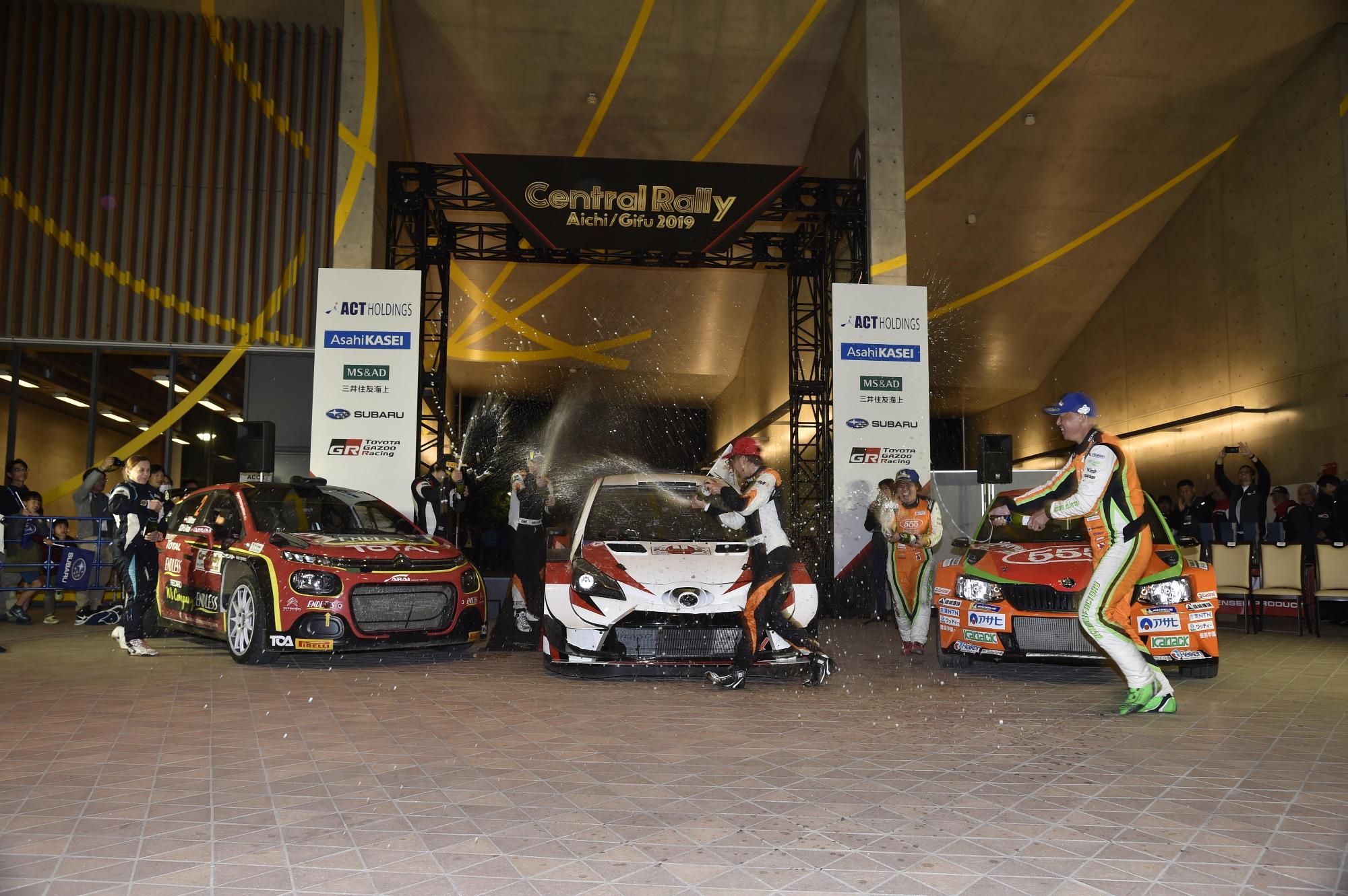 ラリーカー(車)が3台展示されている会場にラリーの選手が集まって、瓶を持ってシャンパンシャワーをかけあっている写真
