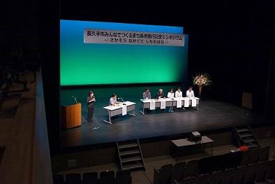 舞台の上の5人のパネリスト、コーディネーター、手話通訳の女性の写真