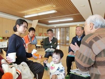 輪になって椅子に腰かけて話し合いをしている小さな子供を連れた母親と4人の男性の写真