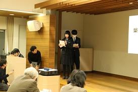 女子生徒と男子生徒が舞台の左側に上がって発表をしており、女子生徒が手に持った資料に目を通しながらマイクを持って話をしている写真