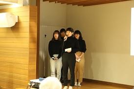 女子学生4名が舞台に上がっており、その中の1名の女子学生がマイクを持って話をしている写真