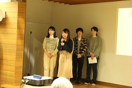 女子学生5名が舞台に上がっており、1名の女子学生がマイクを持って客席の方を見ながら話をしており、後ろの女子生徒が笑顔で見守っている写真