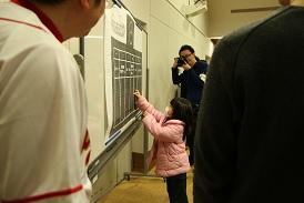 ホワイトボードの前で、ピンク色のダウンコートを着た女の子が投票をしており、それをカメラを持った男性が写真を撮ろうとしており、手前の男性も女の子の投票の様子を見守っている写真