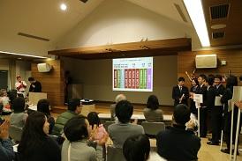 舞台のスクリーンに投票結果が写し出されており、選手の皆さんが自分のチームのプラカードを持って舞台の右横に並んで結果を喜んでおり、観覧の方が拍手をしている写真