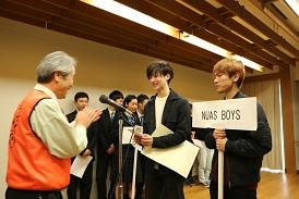 舞台上で表彰が行われており、NUAS BOYSの2人が市長と向かい合って立っており、賞状と商品を受け取って、市長が笑顔で拍手をしている写真