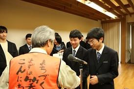 舞台上で、表彰を受けた栄徳高校生徒会チームの男子生徒2名が市長と向かい合って立っており、市長と右側の男子生徒が握手をしている写真