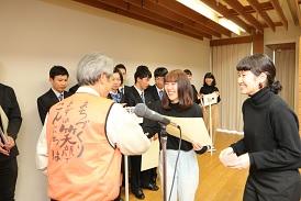 舞台上で表彰が行われており、BABYのチームの中の女子学生2人が市長と向かい合って立っており、賞状を受け取った左側の女子学生が、市長と笑顔で拍手をしている写真