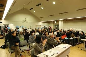 会場の客席にはたくさんのお客さんが見にきて、椅子に座っており、青と白色のボンボンを頭の上で振っていたり、拍手で出迎えている写真