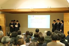 舞台上のスクリーンに資料が映されており、両端に発表の生徒が3名ずつ立っており、左側の男子生徒の1名がマイクを持って、手に持った資料に目を通しながら話をしている写真