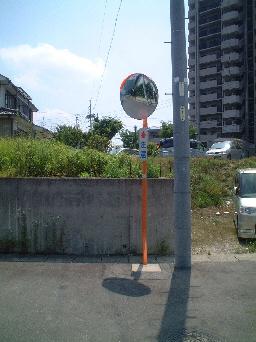 道路に電柱とカーブミラーが並んで設置されており、奥には民家やマンションが写っている写真