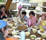 沢山の料理が並んでいる机に参加者がエプロンと三角巾を身につけて「いただきます」をしている写真