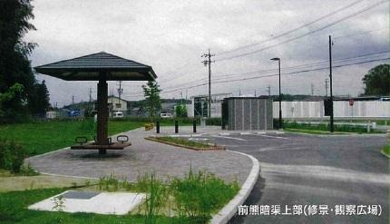 コンクリートなどで舗装してある場所に三角屋根のついた休憩所やレンガで作られた花壇があり、その周辺には緑草が生えている写真