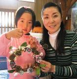 ピンク色の洋服を着た女の子と女性が手作りのクリスマスリースを持って笑顔で写っている写真