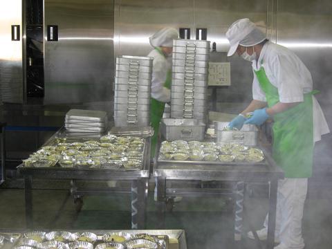 白色の作業着に帽子を被り緑色のエプロンを着たスタッフが、出来上がった蒸しパンをバットに入れている写真