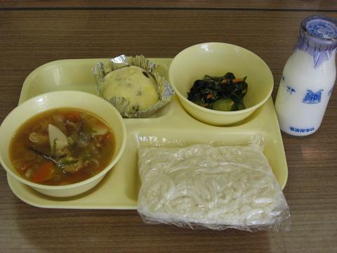 給食のトレーの上に五目うどん、ごま酢あえ、蒸しパンがのせられており、右側に牛乳瓶が置いてある写真