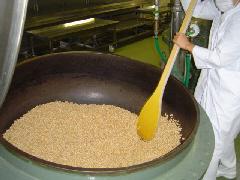 白い作業着を着たスタッフが、大きな釜に材料の大豆と落花生が大量に入っているのを、大きな木べらでかき混ぜている写真